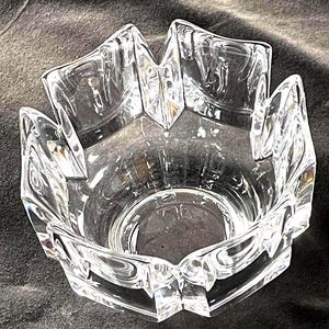 May include: A clear, faceted glass bowl with a starburst design around the rim. The bowl has a hexagonal base and a circular interior. The glass is transparent, allowing light to pass through, and the edges are sharply defined.