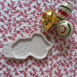 Puede incluir: Un cortador de galletas de cer&aacute;mica blanca con forma de persona sosteniendo una bandera con estrellas. El cortador de galletas est&aacute; sobre un fondo blanco con un patr&oacute;n floral rojo. Hay tres adornos navide&ntilde;os en el fondo: uno dorado, uno rojo y blanco a rayas y uno dorado y verde.