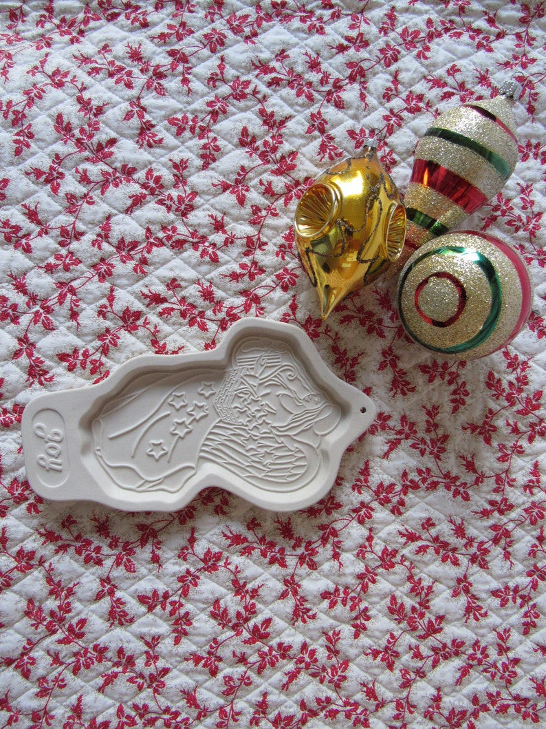 Puede incluir: Un cortador de galletas de cer&aacute;mica blanca con forma de persona sosteniendo una bandera con estrellas. El cortador de galletas est&aacute; sobre un fondo blanco con un patr&oacute;n floral rojo. Hay tres adornos navide&ntilde;os en el fondo: uno dorado, uno rojo y blanco a rayas y uno dorado y verde.