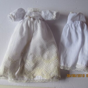 May include: Two white doll dresses, one with lace trim and a ribbon, the other with a simple lace trim.