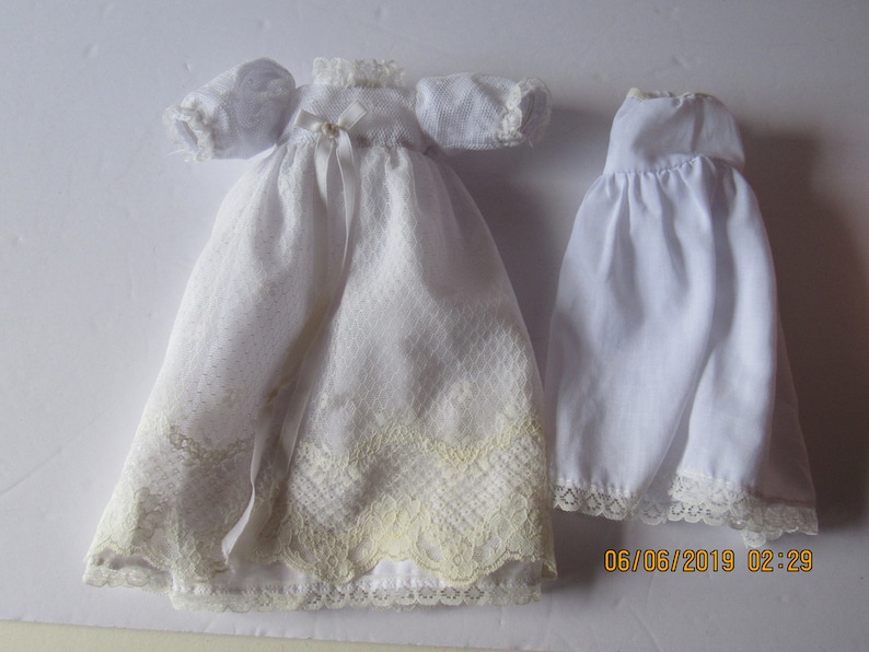 May include: Two white doll dresses, one with lace trim and a ribbon, the other with a simple lace trim.