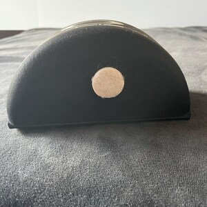 Op de afbeelding: Een zwart, halfrond object met een beige cirkelvormig accent. Het object heeft een gebogen bovenkant en een platte onderkant. Het object is gemaakt van een textuurrijk materiaal.