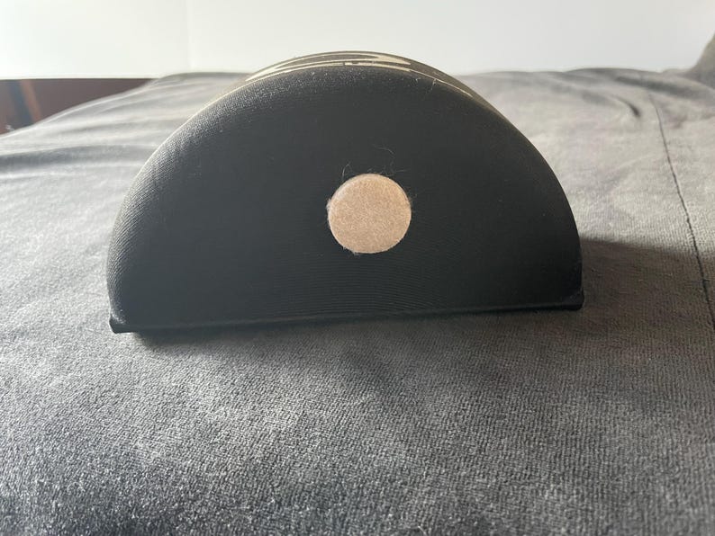 Op de afbeelding: Een zwart, halfrond object met een beige cirkelvormig accent. Het object heeft een gebogen bovenkant en een platte onderkant. Het object is gemaakt van een textuurrijk materiaal.