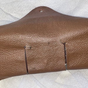 May include: A brown leather eyeglass case with a textured surface. The case has a triangular shape with a small hole at the top and two vertical slits on the front. The case is designed to hold eyeglasses.