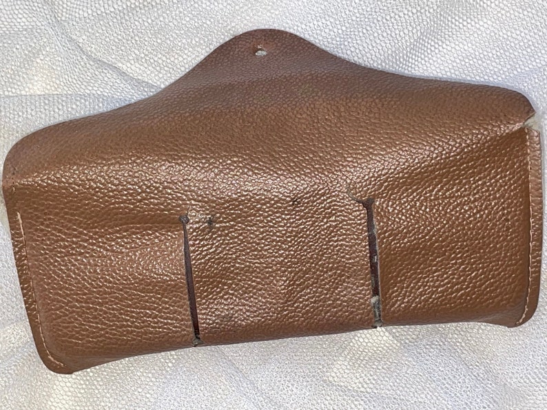 May include: A brown leather eyeglass case with a textured surface. The case has a triangular shape with a small hole at the top and two vertical slits on the front. The case is designed to hold eyeglasses.