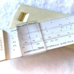 May include: A vintage ARISTO Hydraulic Slide Rule, featuring a clear cursor and white scales with black markings. The slide rule is in a cream-colored case, with measurements in inches.