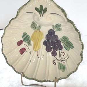 May include: Cream-colored, shell-shaped decorative plate with a scalloped green edge. The plate features hand-painted illustrations of a yellow pear, purple grapes, and red berries with green leaves. The plate is displayed on a gold-colored metal stand.