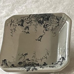 May include: A small, off-white ceramic dish with a square shape and gently curved sides. The dish is decorated with a delicate floral pattern in shades of gray and black, featuring bell-shaped flowers and trailing vines. The dish has a glossy finish.