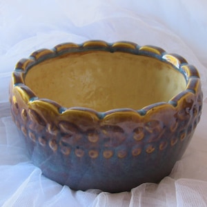 May include: A ceramic planter with a scalloped rim and a textured design of leaves and berries. The planter is glazed in shades of purple, blue, and gold.