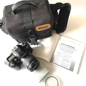 May include: A black Nikon camera kit, including a camera, lens, carrying bag, instruction manual, and lens filter. The camera bag has a brown leather patch with the Nikon logo. The instruction manual is open, displaying text.