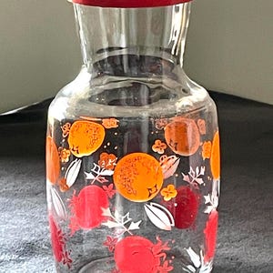 May include: A clear glass pitcher with a red lid, decorated with orange and red fruit designs and white leaf accents. The pitcher is designed for serving beverages and has a wide base and a narrow neck.