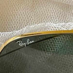 May include: Close-up of Ray-Ban sunglasses. The sunglasses have a gold-colored frame and dark green lenses. The Ray-Ban logo is visible in white on the upper left lens. The background is a soft, white, textured fabric.