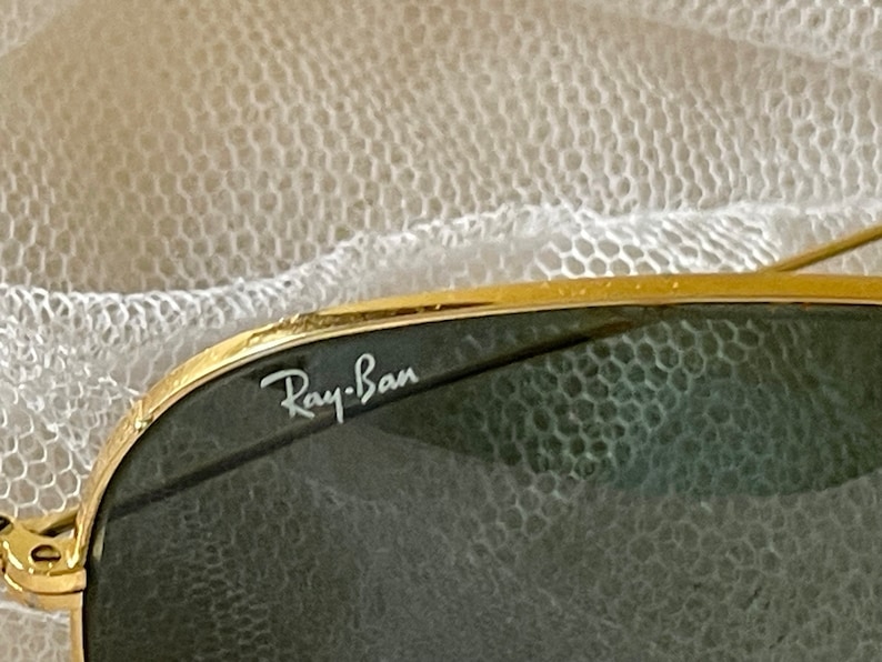 May include: Close-up of Ray-Ban sunglasses. The sunglasses have a gold-colored frame and dark green lenses. The Ray-Ban logo is visible in white on the upper left lens. The background is a soft, white, textured fabric.