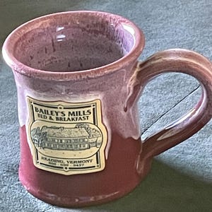 May include: A ceramic mug with a two-tone glaze featuring a light pink top and a dark reddish-brown base. A cream-colored rectangular label on the front displays the text Bailey's Mills Bed & Breakfast, Reading, Vermont, and a phone number.