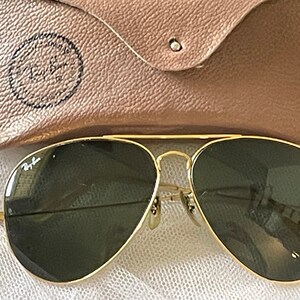 May include: Gold-framed aviator sunglasses with dark green lenses. The sunglasses are next to a tan leather case with the Ray-Ban logo embossed on it. The glasses have a classic design.