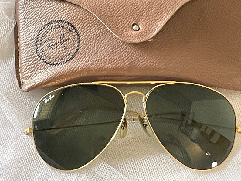 May include: Gold-framed aviator sunglasses with dark green lenses. The sunglasses are next to a tan leather case with the Ray-Ban logo embossed on it. The glasses have a classic design.