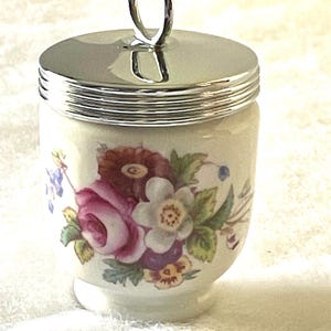May include: A porcelain egg cup with a silver-tone lid and handle. The cup is decorated with a floral pattern featuring pink roses, white flowers, and green leaves. The design is set against a cream-colored background.