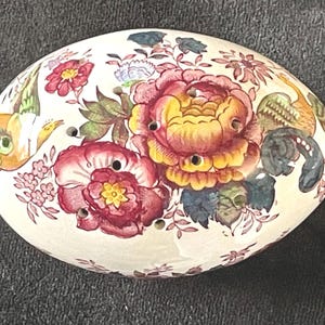 May include: An oval-shaped ceramic flower frog with a cream-colored base. The surface features a colorful floral pattern with large pink, yellow, and red blossoms, green leaves, and small bird illustrations. Several circular holes are spaced across the top.