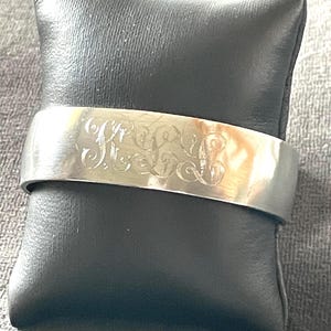 May include: A silver-toned cuff bracelet with an engraved floral design. The bracelet is displayed on a black leather pillow, set against a gray textured background. The cuff is wide and has a polished finish.