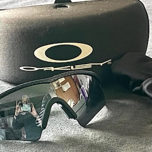 Op de afbeelding: Zwarte Oakley zonnebril met een zwarte etui en schoonmaakdoek. De zonnebril heeft donkere glazen en een zwart montuur. De etui is zwart met het Oakley-logo in wit. De schoonmaakdoek is zwart.