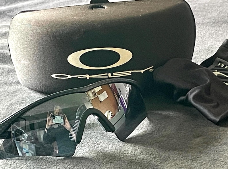 Op de afbeelding: Zwarte Oakley zonnebril met een zwarte etui en schoonmaakdoek. De zonnebril heeft donkere glazen en een zwart montuur. De etui is zwart met het Oakley-logo in wit. De schoonmaakdoek is zwart.