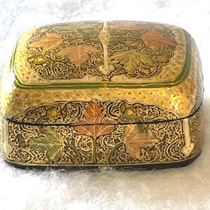 May include: A decorative, rectangular box with a hinged lid. The box is painted with a gold, green, and orange leaf design. The box has a black base and a small, decorative clasp.