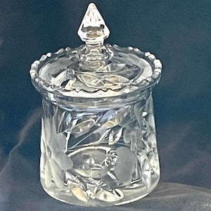 May include: A clear glass jar with a lid. The jar has a round shape with etched floral designs. The lid has a pointed finial and a decorative edge. The jar is likely used for storing small items.