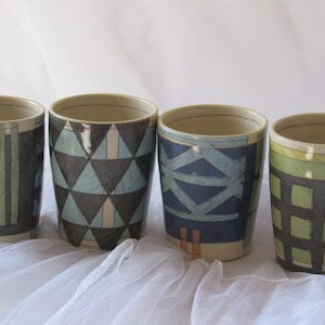 May include: Four ceramic mugs with different geometric patterns. The mugs are in shades of blue, green, black, and white. They are arranged in a row on a white surface.