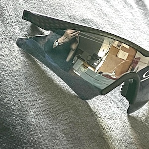 Op de afbeelding: Zwarte en grijze sportzonnebril met een reflecterende lens. De zonnebril heeft een gebogen montuur en een strak, modern design. De lenzen reflecteren de omgeving.