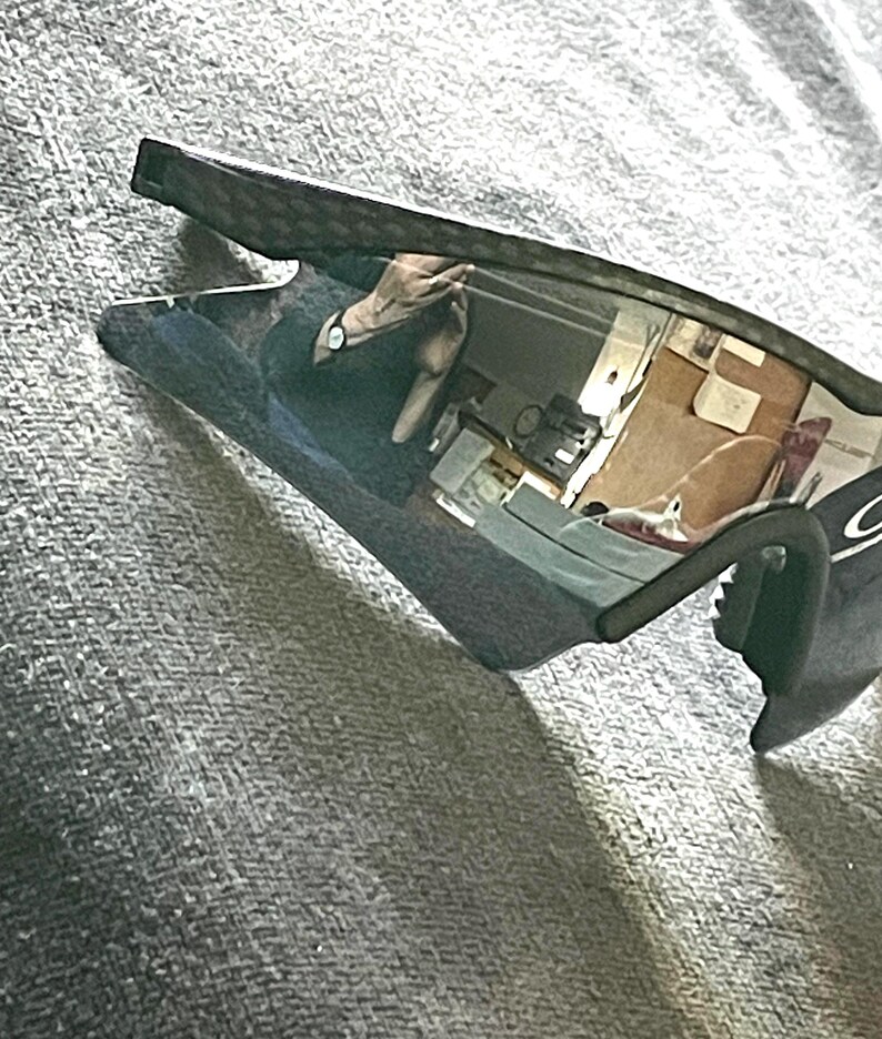 Op de afbeelding: Zwarte en grijze sportzonnebril met een reflecterende lens. De zonnebril heeft een gebogen montuur en een strak, modern design. De lenzen reflecteren de omgeving.