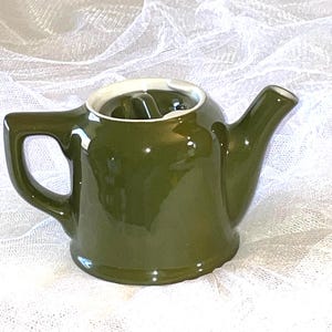 May include: A small, green ceramic teapot with a white interior and a white rim. The teapot has a rounded body and a spout on the side.