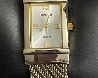 May include: A gold-toned wristwatch with a rectangular face and mesh band. The watch face is white with gold numerals and hands, and the word "RUMOURS" is printed above the hands. The watch is labeled "QUARTZ".