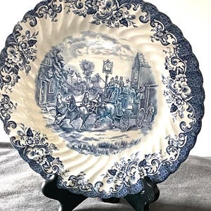 May include: A decorative plate with a blue and white transferware design. The center features a horse-drawn carriage scene, surrounded by a floral border. The plate is displayed on a black stand, showcasing its intricate details and vintage aesthetic.