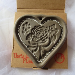 May include: A gray heart-shaped cookie cutter with a raised design of a rose and leaves. The cutter has a white trim around the edge and is in its original packaging. The packaging is brown cardboard with the text "Harts of Flour" printed on it.