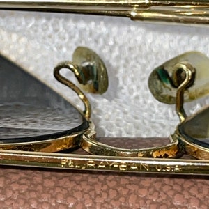 May include: Close-up of gold-framed sunglasses with dark lenses. The frame has a classic design with a bridge and temples. The text "RAY-BAN U.S.A." is visible on the frame. The nose pads are a light beige color.