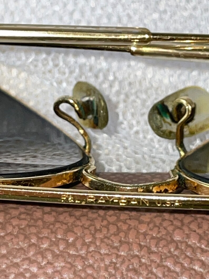 May include: Close-up of gold-framed sunglasses with dark lenses. The frame has a classic design with a bridge and temples. The text "RAY-BAN U.S.A." is visible on the frame. The nose pads are a light beige color.