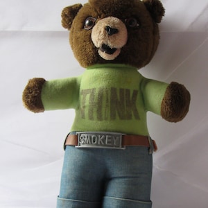 May include: A brown plush bear wearing a green shirt with the word "THINK" printed on it. The bear is also wearing blue jeans and a brown belt with a silver buckle that says "SMOKEY".
