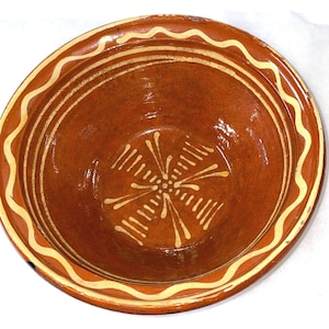 May include: A round, earthenware bowl with a reddish-brown interior and a cream-colored wavy border. The bowl's interior features a cream-colored floral design. The bowl is likely used for serving food.