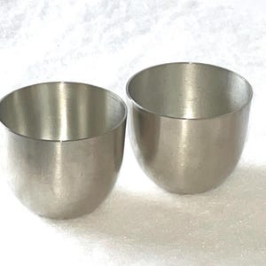 May include: Two silver cups, possibly made of pewter, are shown on a white surface. The cups are round and have a smooth, shiny finish.