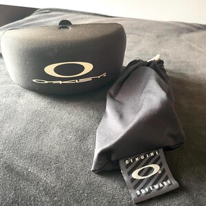 Op de afbeelding: Zwarte zonnebril etui en zacht zakje met het Oakley logo in wit. De etui is ovaal van vorm met een klein lipje aan de bovenkant. Het zakje heeft een label met de tekst "GENUINE SOFTWARE" met het Oakley logo.