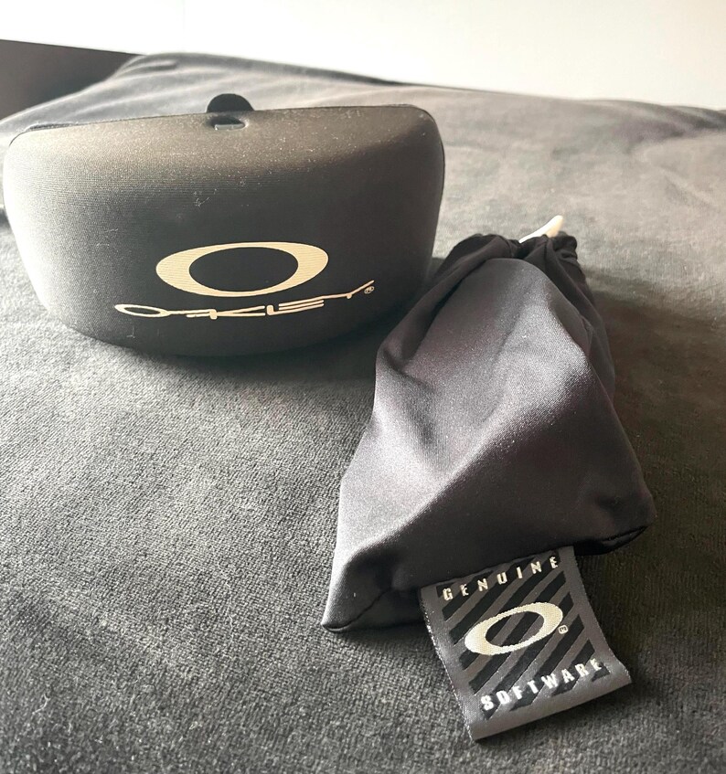 Op de afbeelding: Zwarte zonnebril etui en zacht zakje met het Oakley logo in wit. De etui is ovaal van vorm met een klein lipje aan de bovenkant. Het zakje heeft een label met de tekst "GENUINE SOFTWARE" met het Oakley logo.