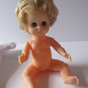 May include: A plastic doll with blonde hair and blue eyes, sitting on a white surface. The doll is wearing no clothes.