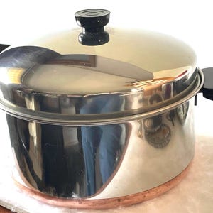 May include: Stainless steel steamer pot with a copper-colored base and black handles. The pot has a shiny, reflective surface and a matching lid with a black knob. The pot is designed for steaming food.