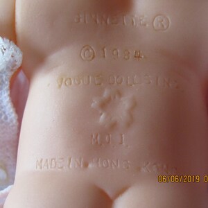 May include: Close-up of a doll's back with a white lace dress. The doll has a label that reads "Vogue Dolls Inc." and "Made in Hong Kong" with a copyright symbol and the year 1984.