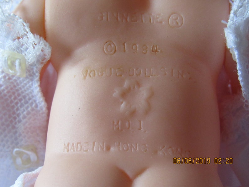 May include: Close-up of a doll's back with a white lace dress. The doll has a label that reads "Vogue Dolls Inc." and "Made in Hong Kong" with a copyright symbol and the year 1984.