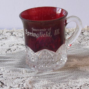 May include: A red glass souvenir mug with a clear glass handle. The mug has a cut glass design at the bottom and says "Souvenir of Springfield, Ill." on the side.
