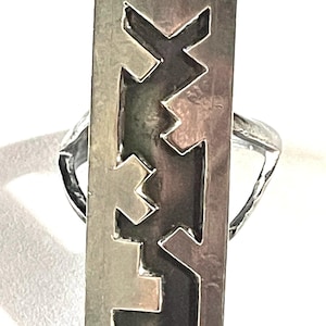 May include: A silver ring featuring a tall, rectangular face with a cutout geometric pattern. The metal surface has a polished finish, while the interior of the cutout design appears dark, creating a high-contrast visual effect on the band.