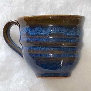 May include: Handmade ceramic mug with a dark brown rim and handle, transitioning to a deep blue body. The mug features horizontal textured lines and a rounded shape, perfect for coffee or tea.