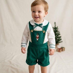 May include: A white long-sleeved shirt with a green collar and cuffs embroidered with the name Gabriel, paired with green overalls featuring a nutcracker graphic, gold buttons, and a small green bow at the neckline.