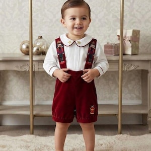 Puede incluir: Una camisa blanca de manga larga con cuello redondo y bordados de estrellas doradas, combinada con pantalones cortos de terciopelo rojo oscuro con tirantes. Los tirantes muestran figuras de cascanueces y el pantalón tiene un tambor bordado.